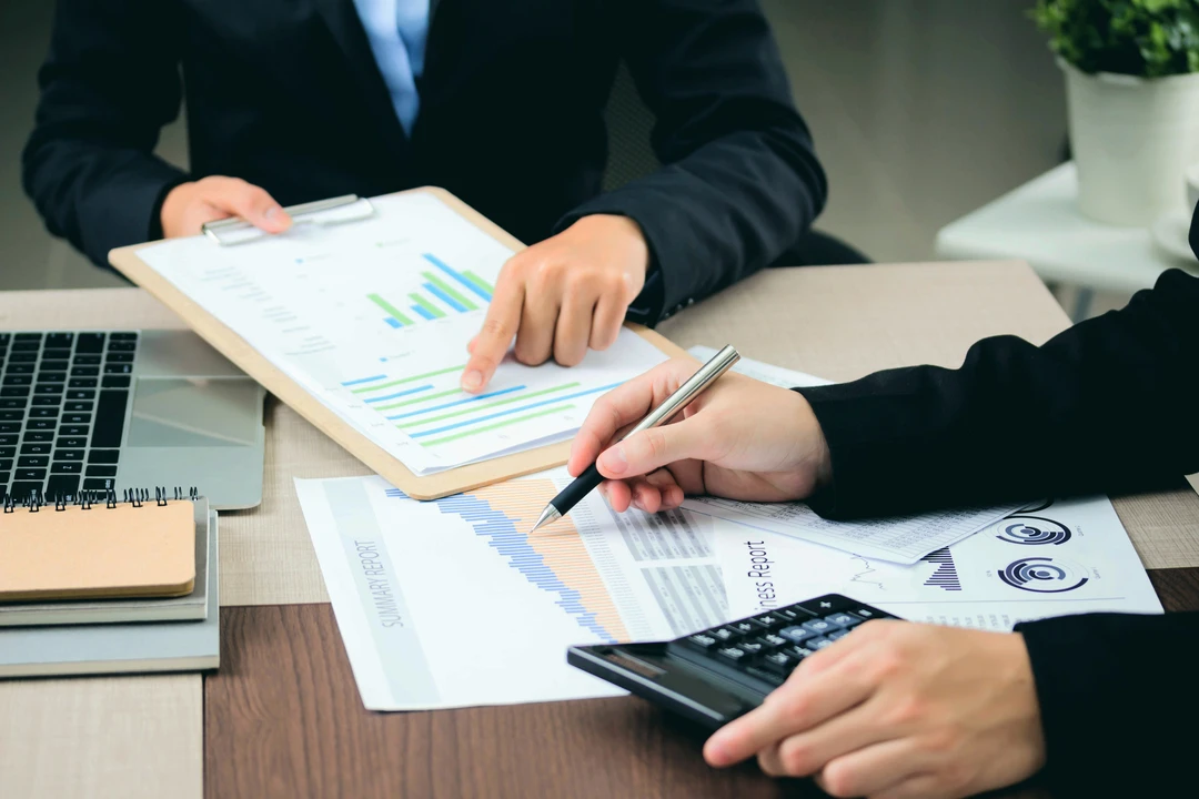 Two professionals reviewing business charts and reports at a desk, with a laptop and calculator present.