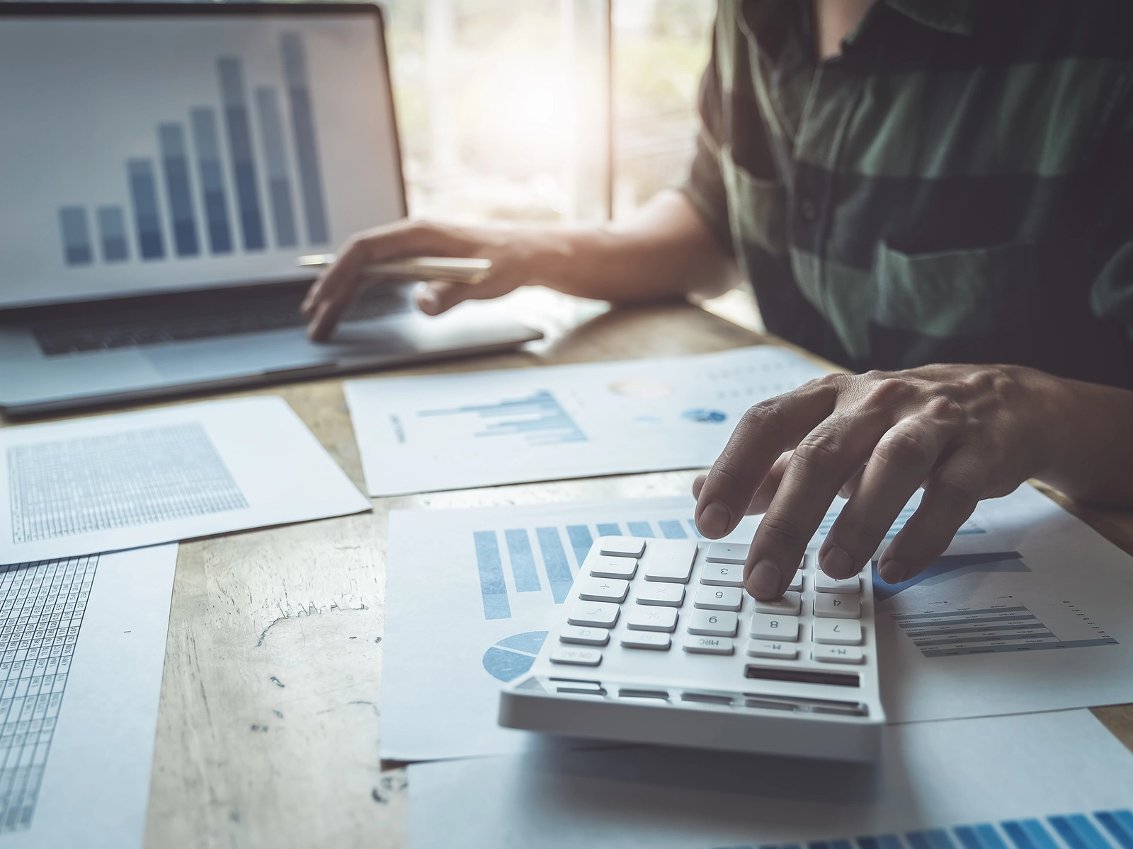 A person using a calculator while working on a laptop, surrounded by printed graphs and charts on a wooden table.