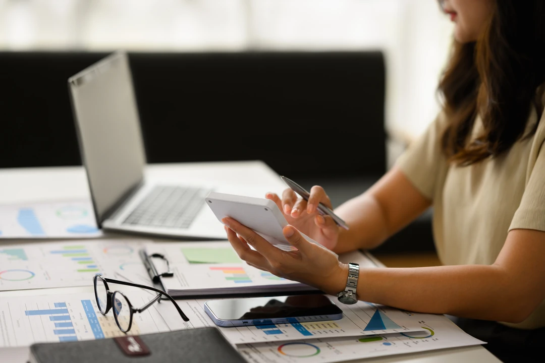 A person holds a tablet and pen while reviewing charts and graphs on a desk with a laptop and glasses nearby.