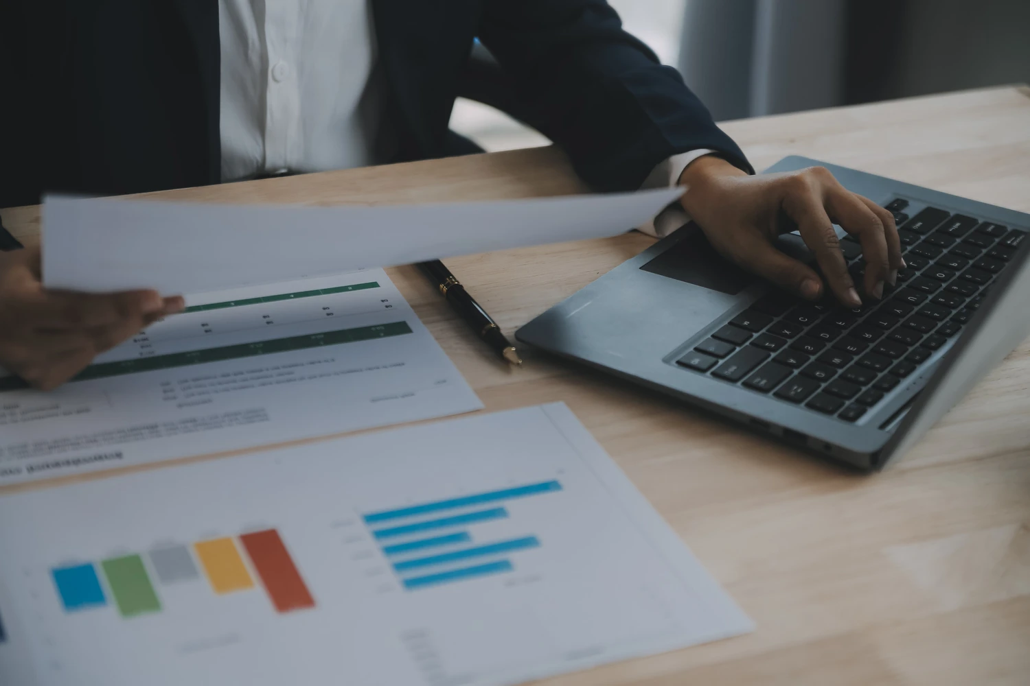217379717_m A person in a suit reviewing documents with graphs and charts while typing on a laptop at a wooden desk.