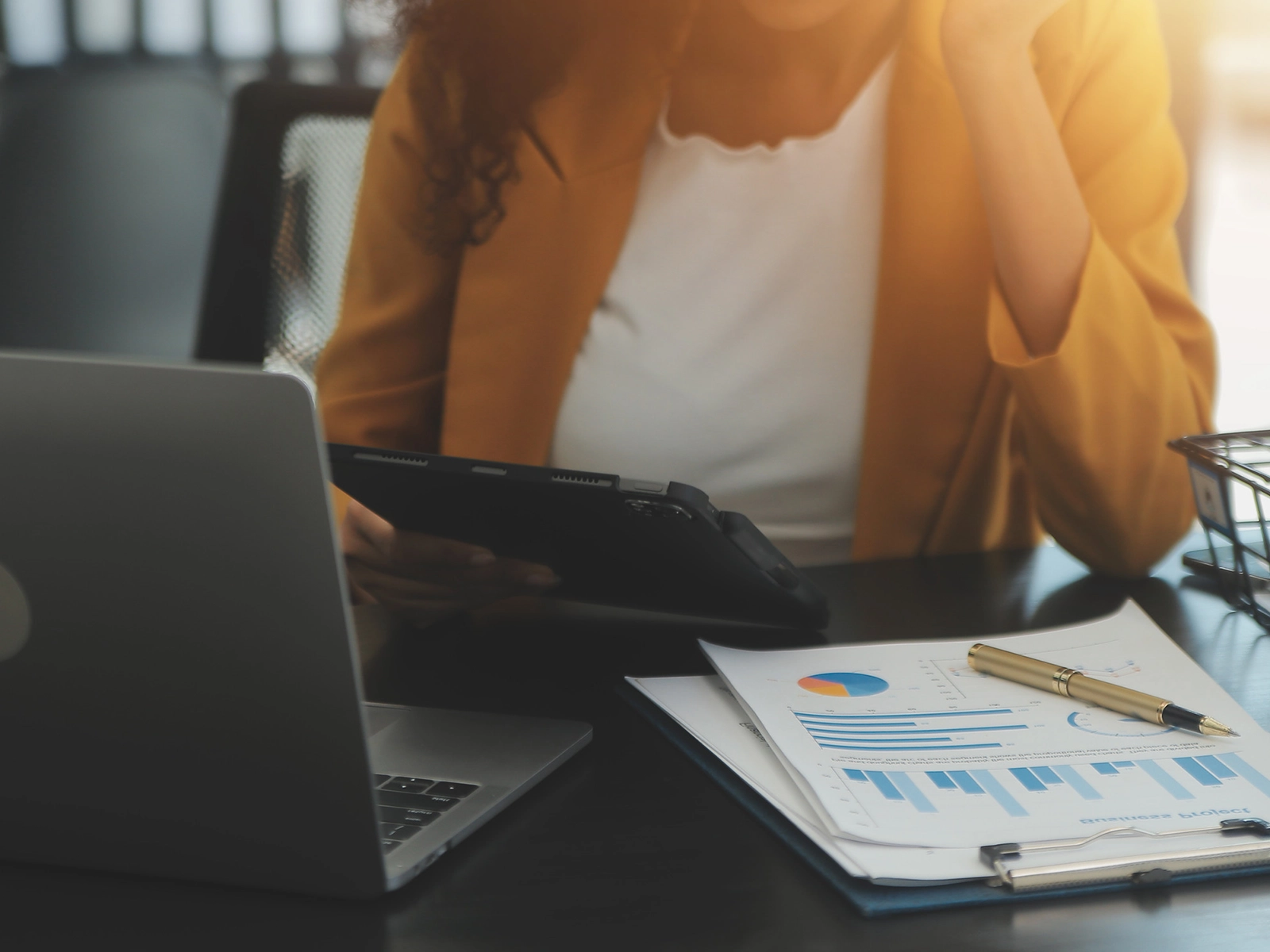A person in a yellow blazer is analyzing data on a tablet at a desk, with a laptop and charts nearby, showcasing a professional workspace.