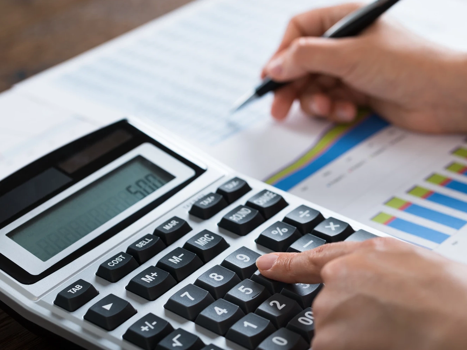 Close-up of hands using a calculator and a pen to work on financial documents containing colorful bar charts.